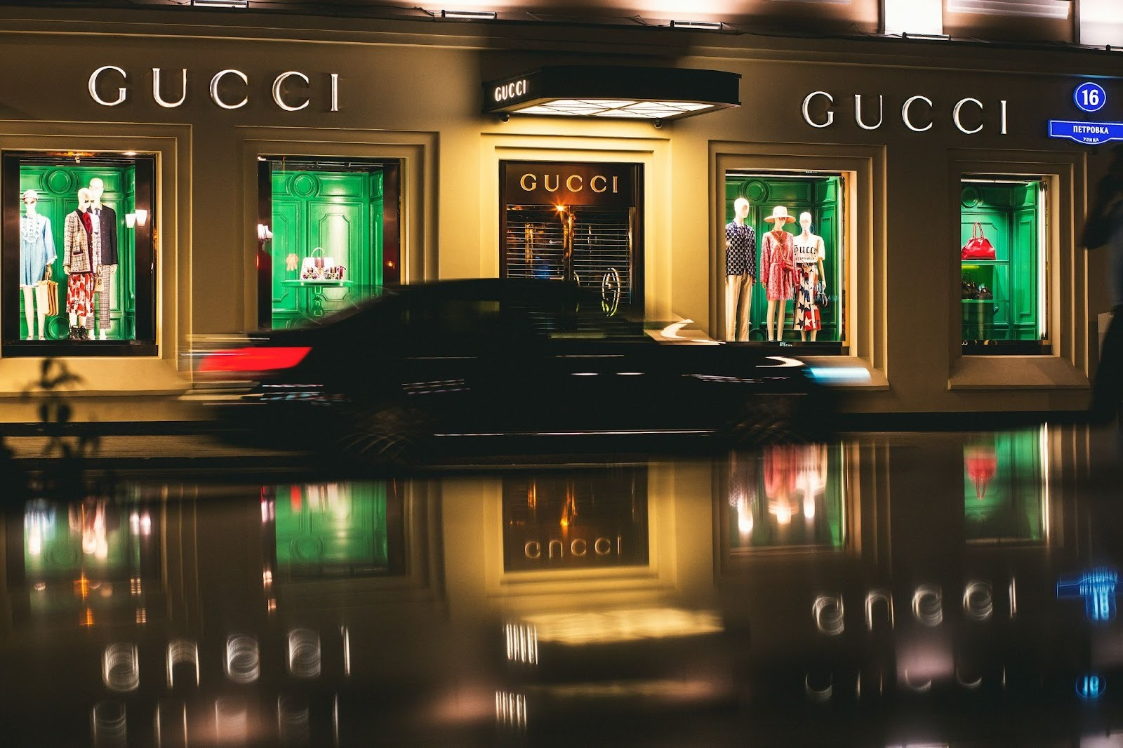 Car drives by Gucci store with window displays of fashionable clothing.