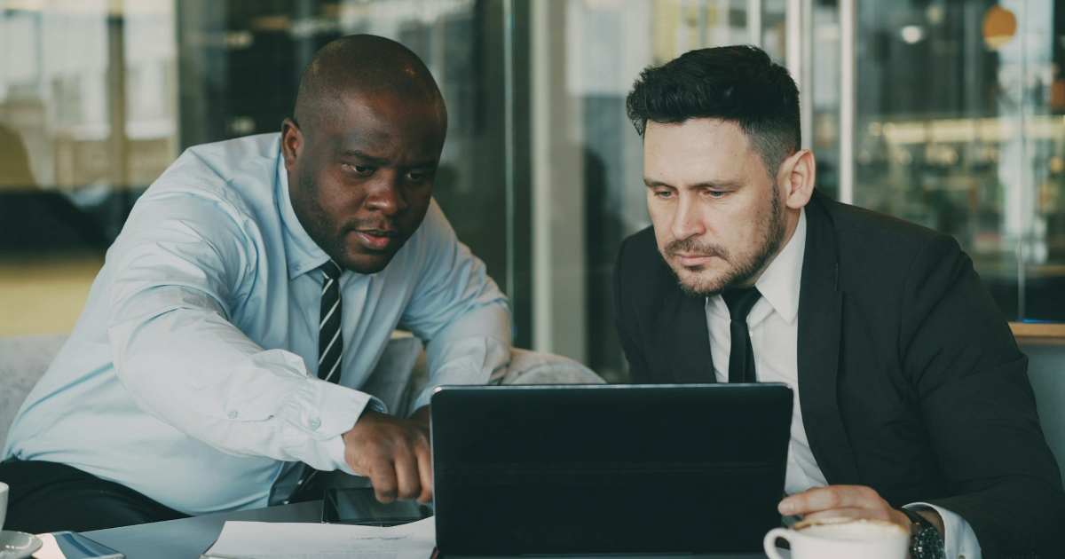 Two male coworkers look at a laptop together, with one pointing at the screen