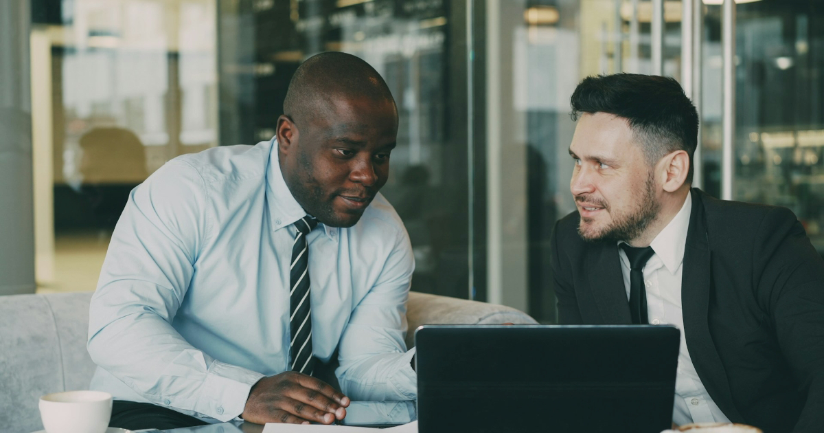 One male coworker looks to another as they sit in adjacent chairs and look at a laptop together