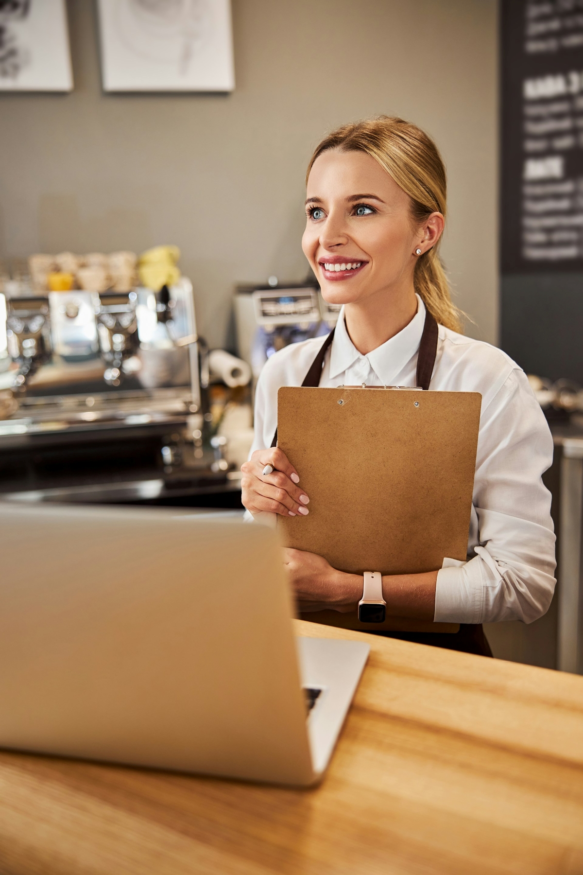 A female hospitality worker looks happy while holding a clipboard behind a counter with a laptop on it and in front of a coffee machine