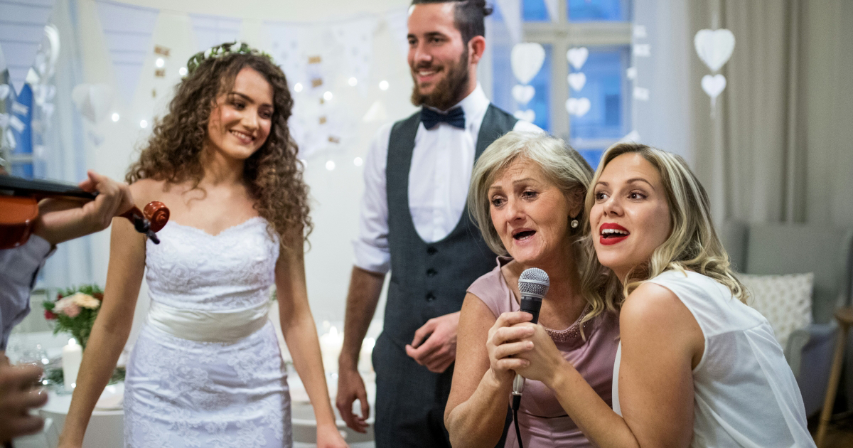 An older female wedding guest sings karaoke with a younger woman as the bride and groom look on
