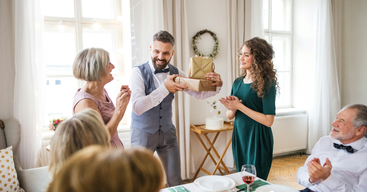 A groom stands at the head of a table, holding gifts, his mom and his fiancée standing either side of him 