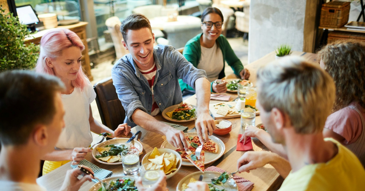 Friends eating dinner at a restaurant together, with one man putting a slice of pizza on another's plate