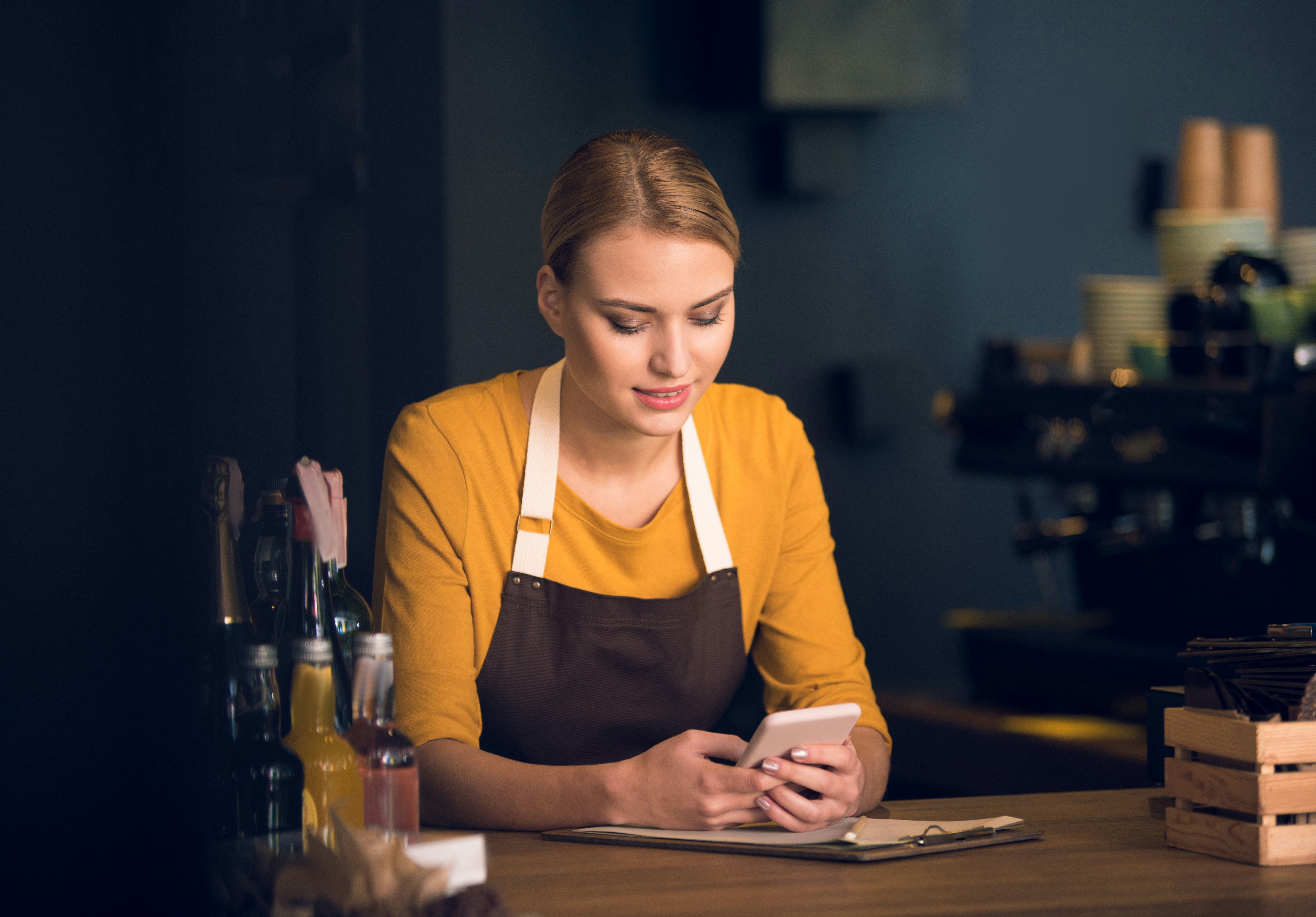 Coffee shop employee looks at her phone, full of text messages from her boss, as a model depicts. 