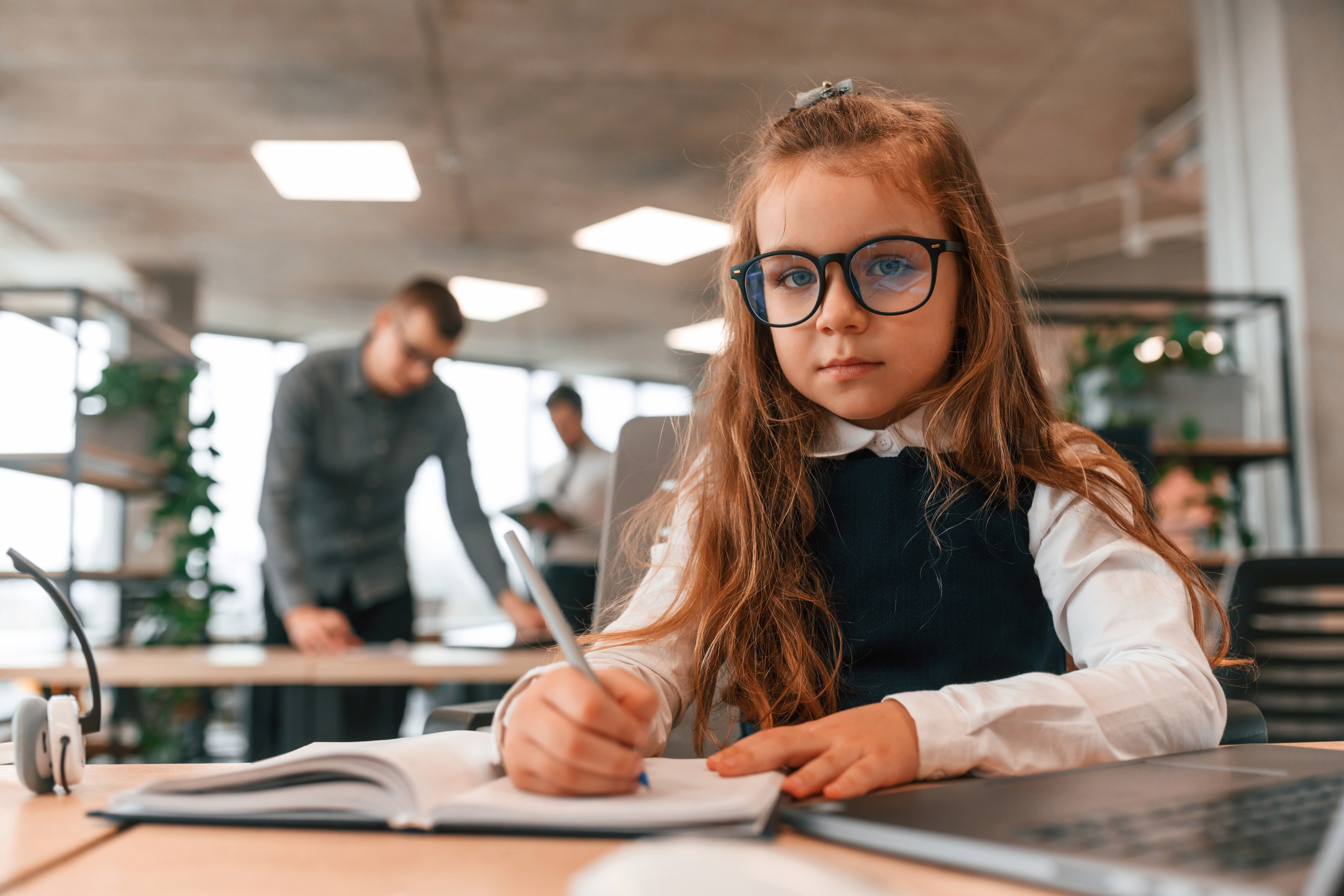 Young girl wearing glasses writes in a notebook at an office desk with adults in the background.