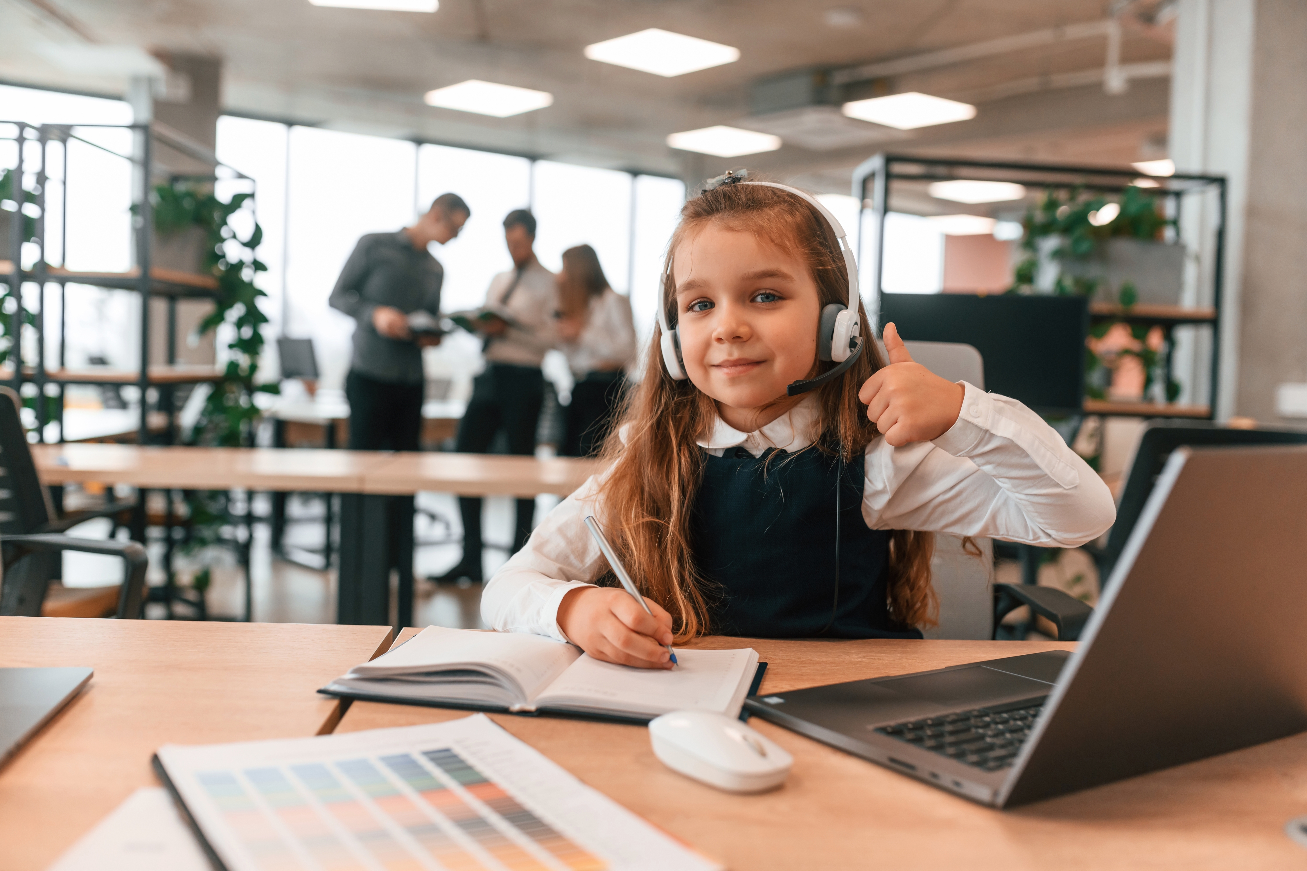 Young child wearing a headset at an office desk with a laptop and notebook.