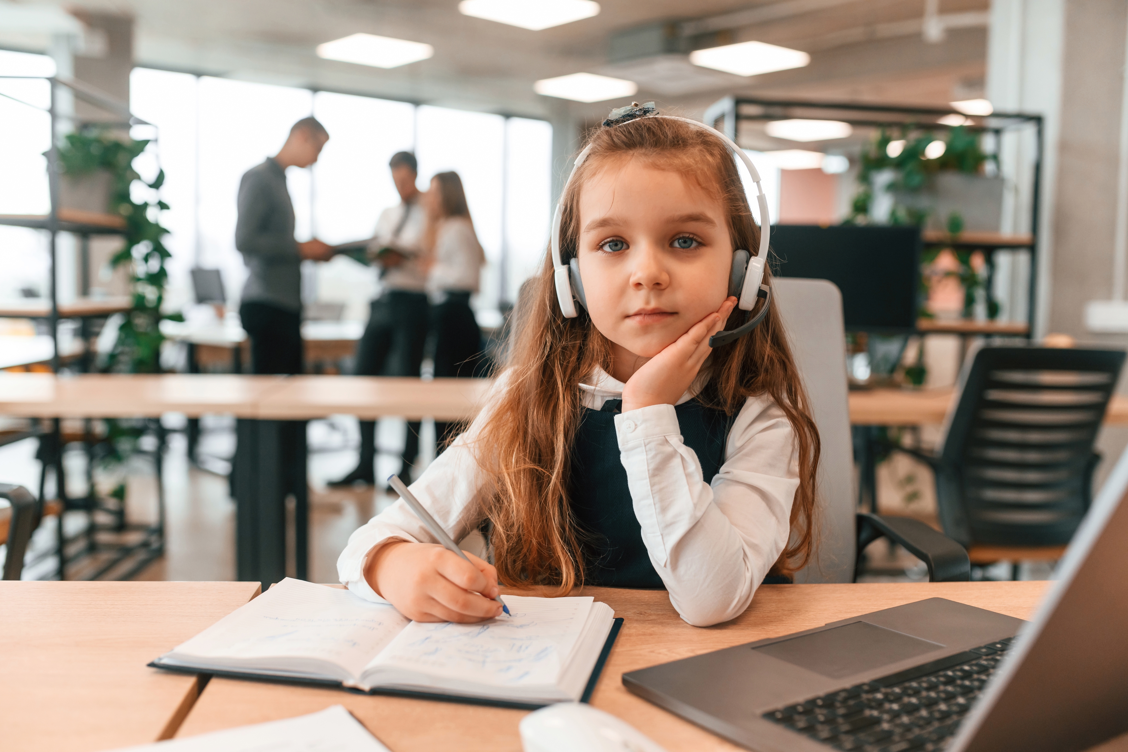 Young girl wearing a headset sits at an office desk with a notebook and laptop.