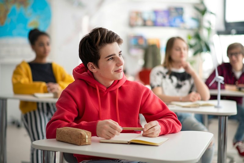 A high school student in a red sweater sits at his desk with a mischievous smile.
