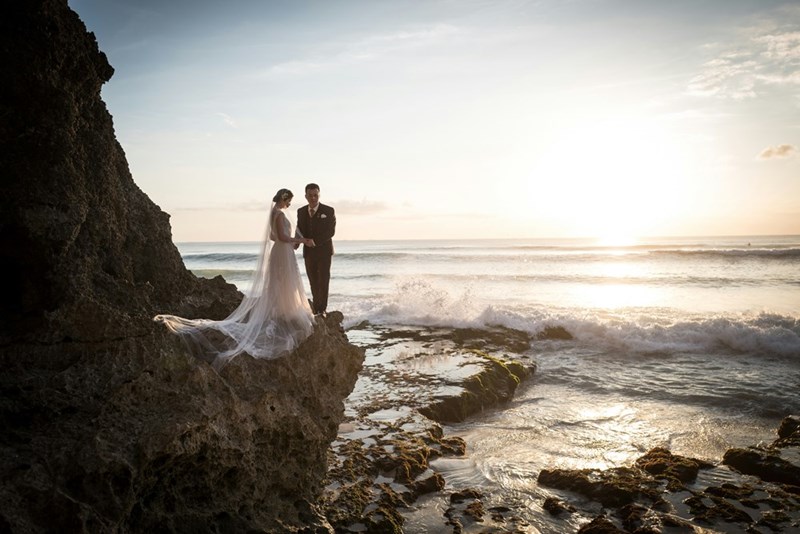 A bride and groom standing on a rocky cliff overlooking the ocean