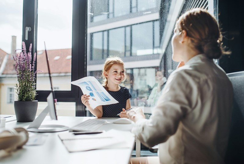 Worker sits in office with her smiling daughter, who holds a paper.