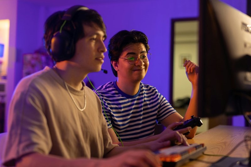 A gamer plays on his computer as his roommate looks on.