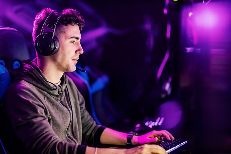 Young male gamer wearing a headset plays a video game on a desktop computer in a neon-lit gaming setup, focused on the screen with purple and blue LED lighting.