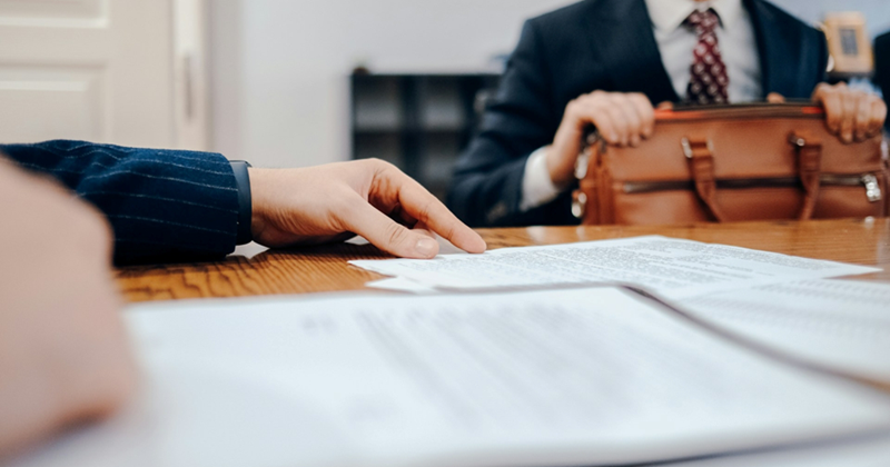 A man in a suit holding a briefcase sits across from another person at a table with papers on it