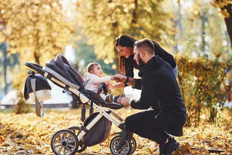 Dad holds baby's hand while Mom looks on as they kneel next to a stroller in the park.
