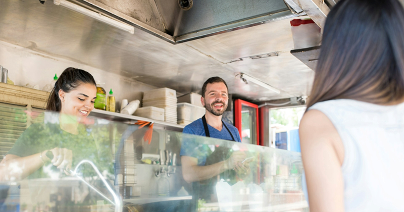 A man serves a customer at a food truck while a woman cooks beside him
