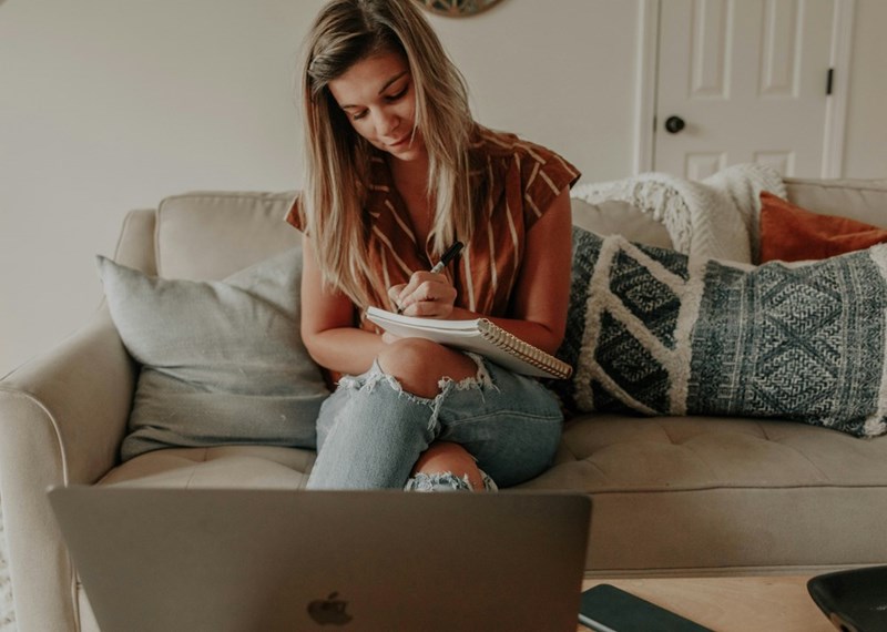 Young woman sitting on a couch in a cozy living room, writing in a spiral notebook while looking toward a laptop on a coffee table, surrounded by decorative pillows.