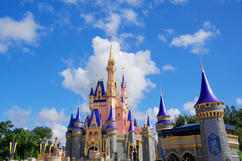 Cinderella Castle at Walt Disney World with pink walls and blue spires under a bright blue sky with scattered clouds, surrounded by stone towers and decorative banners.