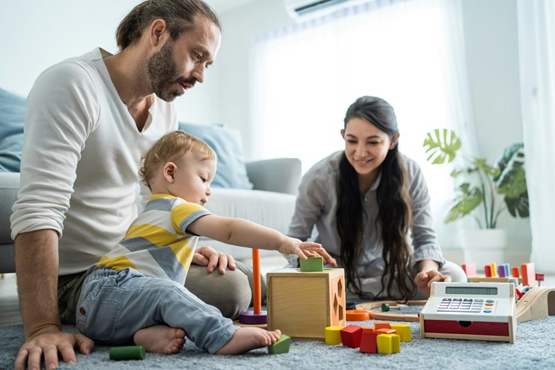 2-year-old sits next to father playing with toys while Mom looks on.