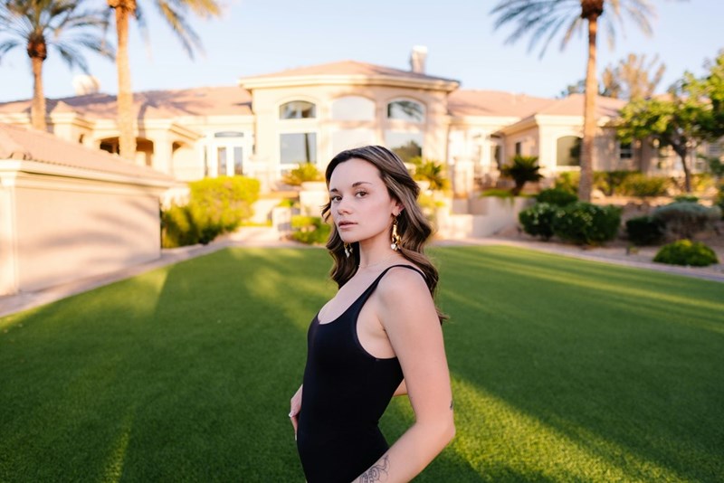 Wealthy teen college girl stands proudly in front of large home with perfect lawn.