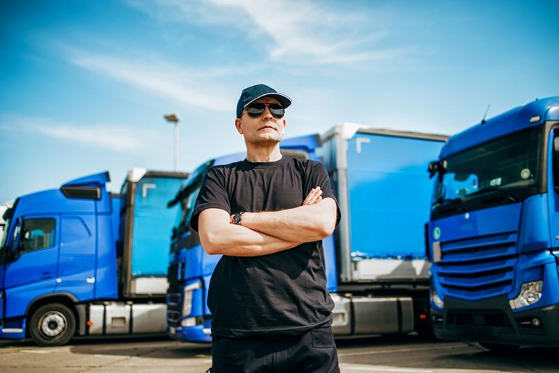 Professional truck driver with hat and sunglasses confidently standing in front of big and modern truck