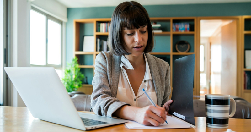 An office worker sitting at a desk makes notes in a folder with an open laptop on the desk in front of her