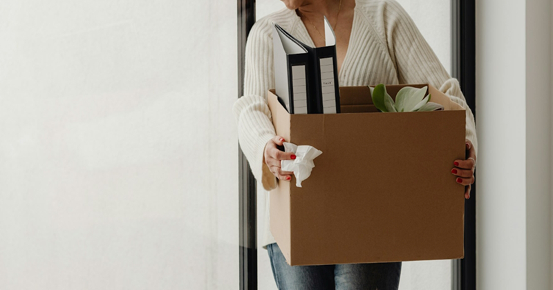 A woman holding a cardboard box of belongings walks through a doorway