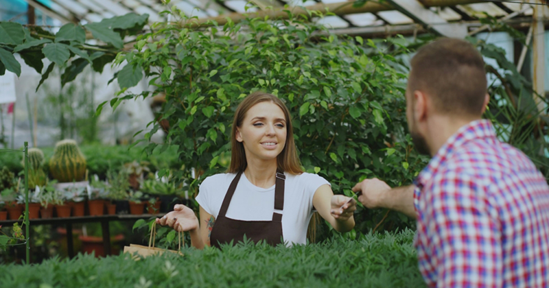 A woman working in a greenhouse reaches across a table full of plants to give a male customer a cutting