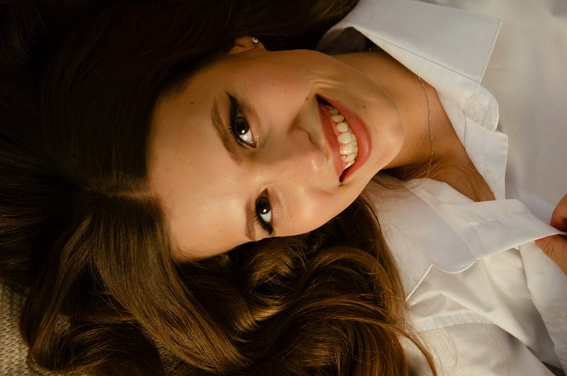 Close-up portrait of a smiling young woman with long brown hair lying down, wearing a white shirt and looking up toward the camera in warm natural light.