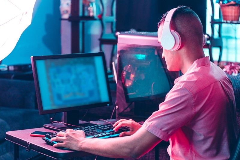 Young man wearing white headphones gaming on a desktop computer with dual monitors in a neon-lit room, using a keyboard and mouse at a small desk setup.