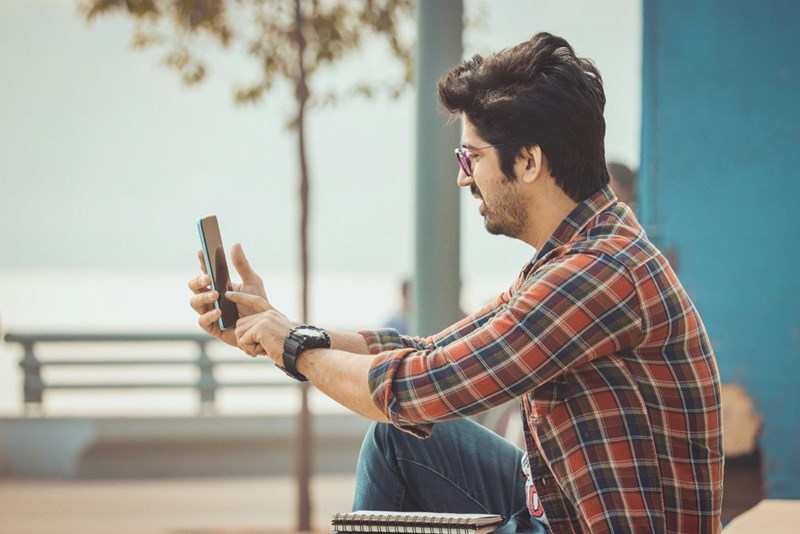 Man wearing glasses and a plaid shirt sitting outdoors, holding a smartphone in both hands and taking a photo or recording a video near a waterfront promenade.