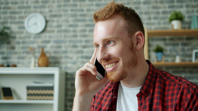 Smiling red-haired man with a beard talking on a smartphone in a modern home office with a brick wall, shelves, and a clock in the background.