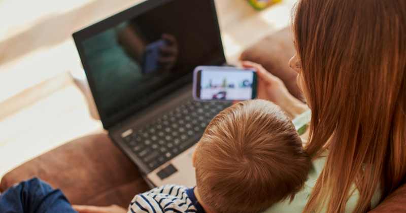 A mom sitting on a couch holds her phone up for the child beside her to watch while working on her laptop