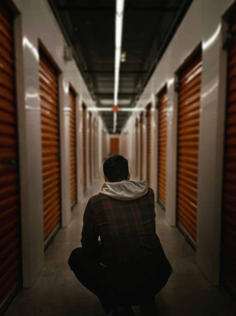 Person sitting alone in a narrow hallway of indoor storage units, facing away from the camera, surrounded by rows of orange roll-up doors under fluorescent lighting.