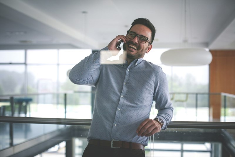 Smiling businessman wearing glasses and a button-down shirt talking on a smartphone while leaning on a railing inside a modern office building with large windows.