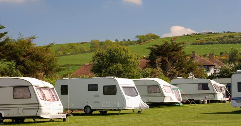 Row of white travel caravans parked on a grassy campsite with rolling green hills and countryside homes in the background under a partly cloudy sky.