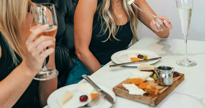 Two women drink out of wine glasses and share a charcuterie board in a restaurant