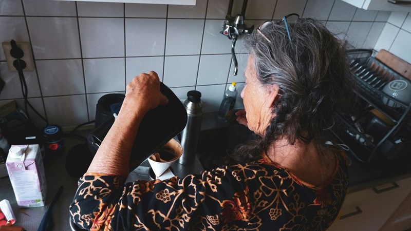 An older grandparent makes coffee in the kitchen while their back is turned away from the dinner table.