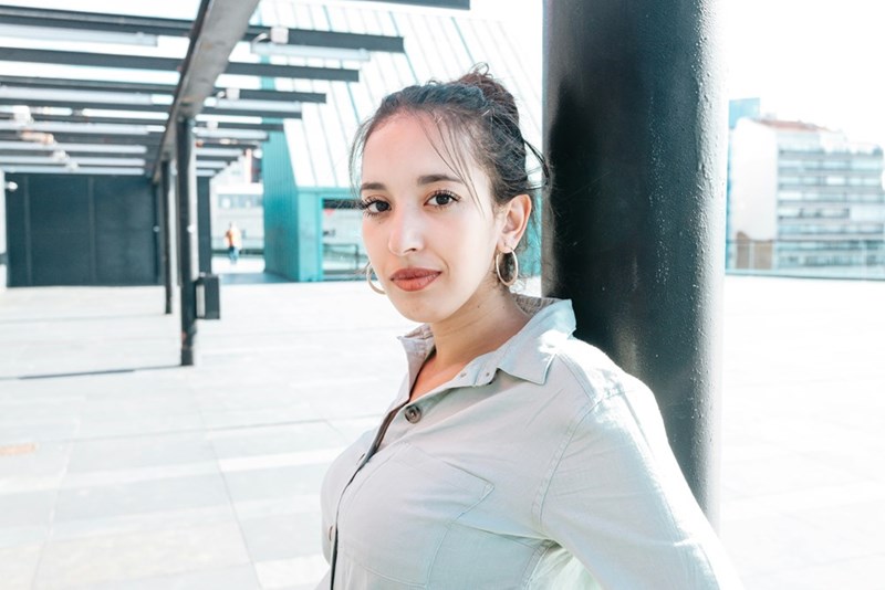 Young woman with her hair in a bun and hoop earrings leaning against a black pillar on a modern rooftop terrace, looking directly at the camera.