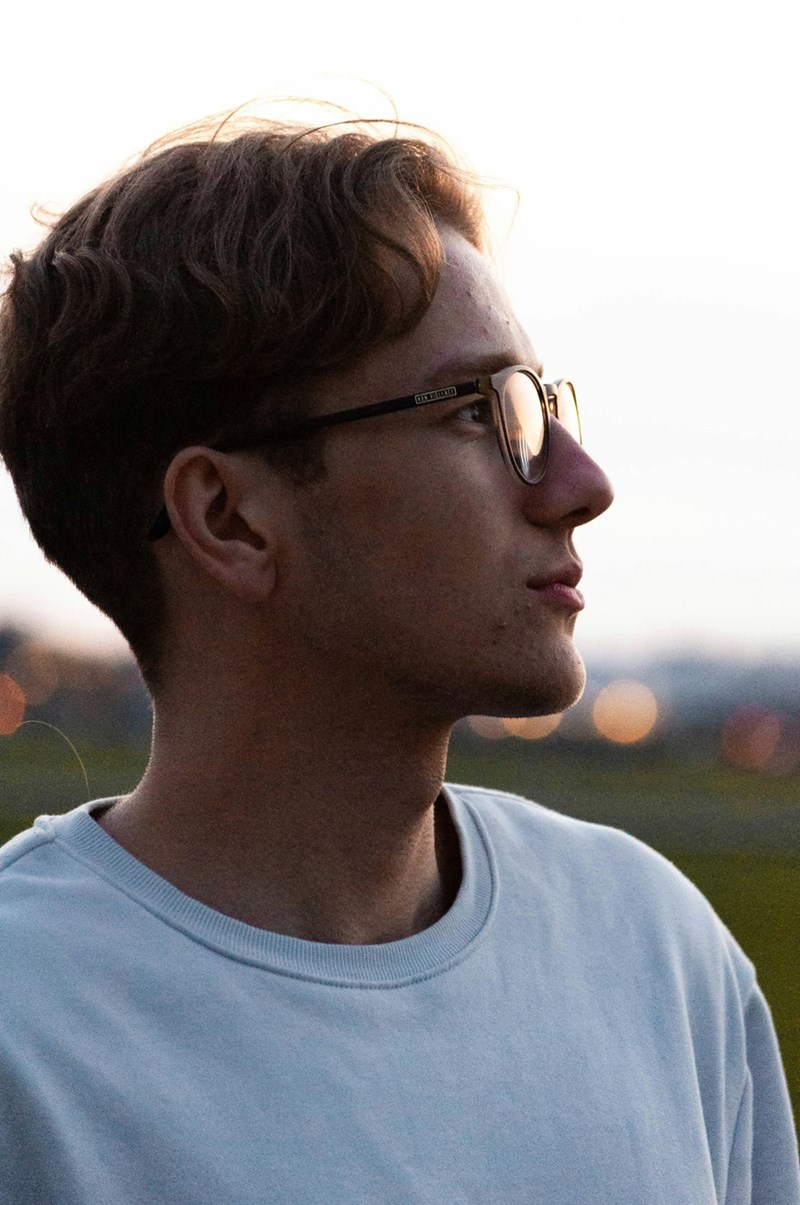 Side profile of a young man wearing glasses and a light sweatshirt, looking into the distance at sunset with soft city lights blurred in the background.