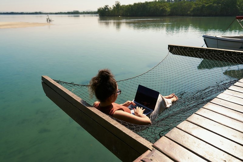 Relaxed employee works from hammock over water in bright sunlight.