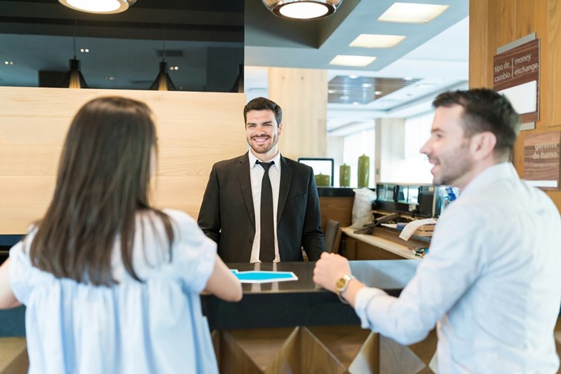 Two customers check in at the front desk with a male receptionist.