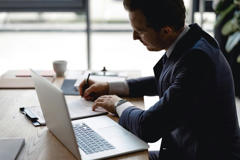 Businessman in a dark suit sitting at a table with his laptop, signing a document