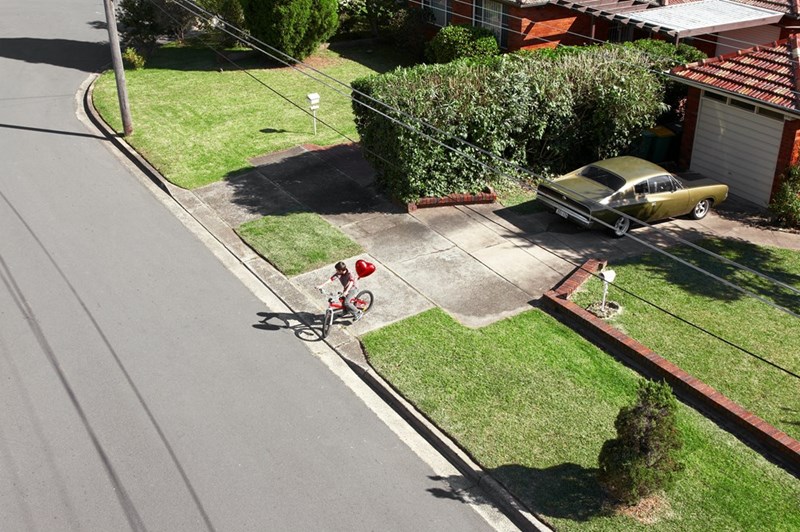 Aerial view of a child riding a bicycle along a quiet suburban street with a red heart-shaped balloon tied to the bike near a driveway and parked car.