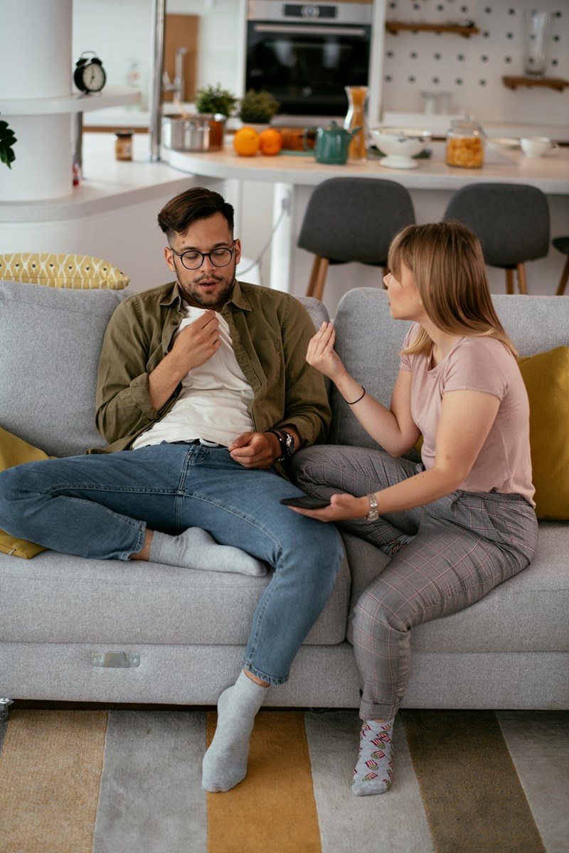 Couple sitting on a living room couch having a serious conversation, with the woman gesturing while the man looks concerned in a modern home interior.
