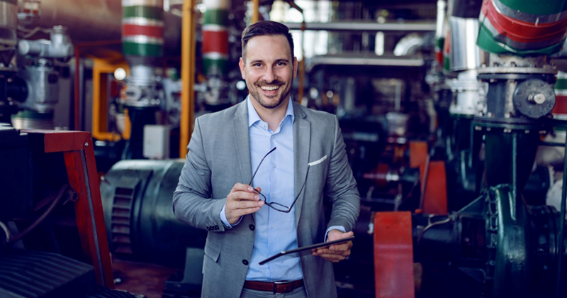 A businessman holding a clipboard smiles while standing on a manufacturing floor