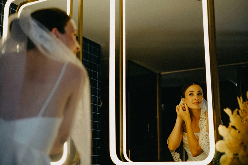 The bride adjusts her earring while getting ready in front of a mirror.