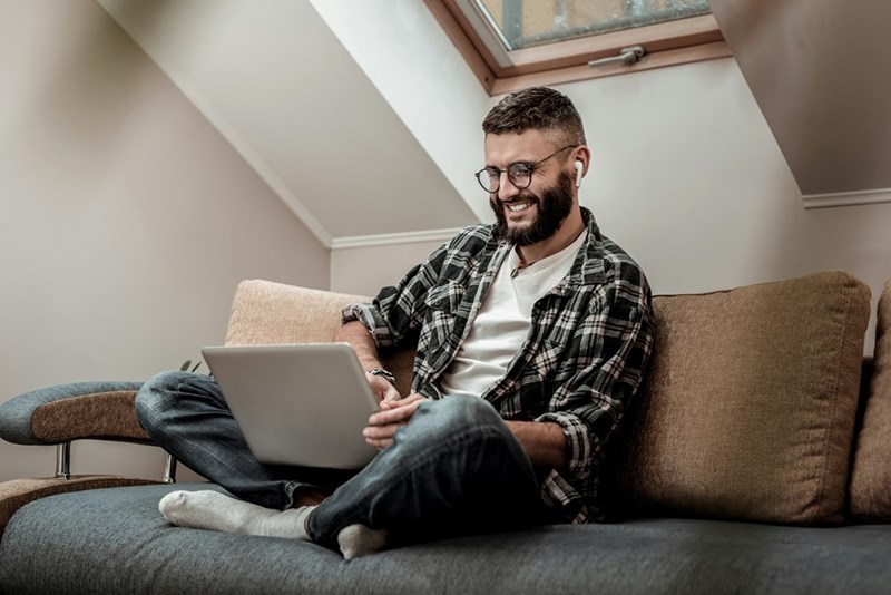Bearded man wearing glasses and earbuds sitting cross-legged on a couch under a skylight, smiling while using a laptop at home.