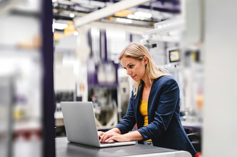 Woman in a blazer working on a laptop inside a modern factory or industrial workspace.