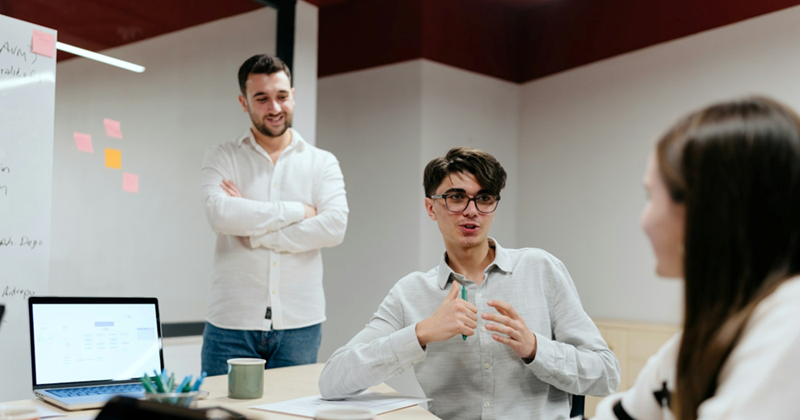 A man stands in the background with his arms crossed as another man, sitting at a table with a laptop, talks to a woman also sitting at the table