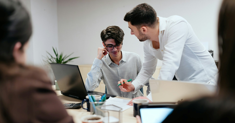 A man stands over another man sitting at a table with his laptop, talking to him, while two other seated people watch
